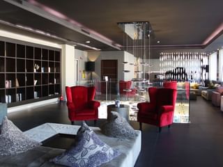 Hall d'entrée moderne C-Hotels avec sols sombres, fauteuils en velours rouge, tables en verre, étagères intégrées et éclairage contemporain.