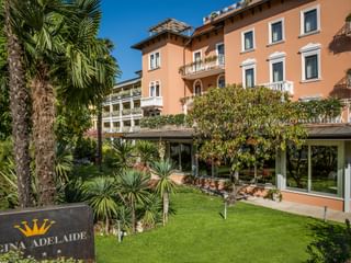 Élégant Hôtel Regina Adelaide de couleur corail avec balcons blancs, entouré de palmiers et jardins entretenus sous ciel bleu.