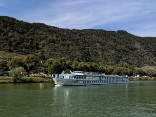 Bateau de croisière MS Switzerland naviguant sur eau verte calme avec collines boisées et petit village en arrière-plan sous ciel bleu.