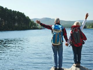 couple de randonneurs qui se tiennent la main de dos en observant devant eux un lac et ses sapins