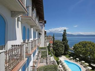 Terrasse de l'Hôtel Savoy Gardone avec volets bleus donnant sur le lac de Garde, avec piscine incurvée entourée de jardins luxuriants.