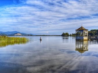 Lac tranquille avec pavillon en bois sur pilotis, roseaux au premier plan, montagnes lointaines sous ciel nuageux près de Viareggio, Italie.