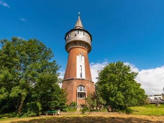 Château d'eau en brique rouge à Husum avec toit pointu et entrée blanche, entouré d'arbres verts et de pelouse sous ciel bleu.
