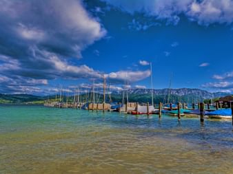 Jetée en bois avec voiliers et bateaux à moteur amarrés au lac Attersee. Eau turquoise claire, ciel nuageux dramatique et montagnes alpines.