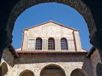 Vue à travers des arcs de pierre de la basilique euphrasienne à Porec, montrant une façade ornée avec trois fenêtres cintrées.