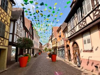 Rue pavée dans un village de la Route des Vins d'Alsace avec maisons à colombages colorées et décorations de feuilles de vigne suspendues.