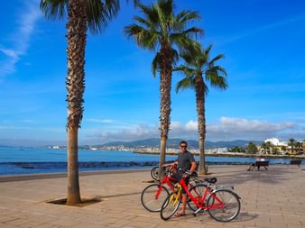 Homme debout avec des vélos rouges sur la promenade du front de mer à Palma, Majorque. Des palmiers bordent la promenade avec mer bleue et montagnes.