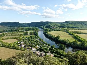 Vue aérienne de la Dordogne serpentant à travers une vallée verdoyante avec champs, forêts et collines. Un pont traverse la rivière.