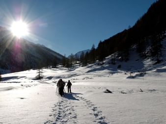 Deux randonneurs en raquettes marchent dans la neige profonde du Queyras avec un soleil éclatant, un ciel bleu et des sommets enneigés.