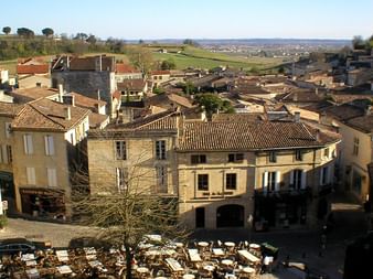 Vue aérienne d'une place de village avec terrasse de café dans le vignoble bordelais. Bâtiments en pierre aux toits de tuiles entourent la place.
