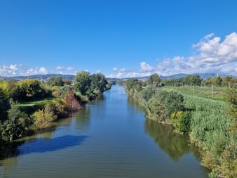 Rivière calme traversant la campagne toscane verdoyante avec des arbres bordant les deux rives, collines lointaines et ciel bleu.