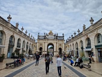 Vue de la Place Stanislas à Nancy montrant l'Arc Héré orné de détails dorés, flanqué de bâtiments symétriques avec arcades.