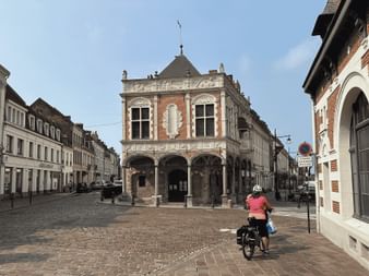 Cycliste avec vélo chargé sur place pavée en Flandre française, face à bâtiment en brique orné avec colonnade à arcades.