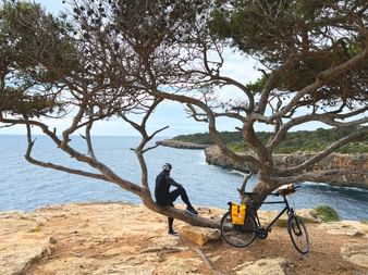 Cycliste en vêtements noirs assis sur une branche de pin surplombant la Méditerranée à Majorque, vélo garé à proximité sur la côte rocheuse.