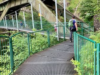 Randonneur avec sac à dos marchant sur passerelle métallique verte avec garde-corps, passant sous un pont en béton entouré de forêt.
