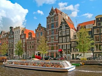 Bateau touristique sur un canal d'Amsterdam passant devant des maisons à pignons néerlandaises avec toits orange-rouge sous ciel bleu.