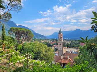 Clocher de Merano avec toile de fond alpine Vue depuis le Tappeinerweg à Merano montrant un clocher historique entouré d'une végétation luxuriante et de montagnes en arrière-plan.