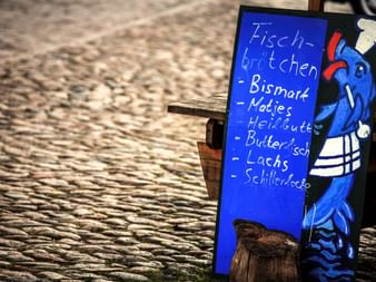 Panneau en bois bleu avec chef poisson cartoon annonçant sandwichs au poisson et spécialités de fruits de mer sur rue pavée.