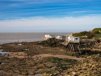 Cabanes de pêche traditionnelles sur pilotis en bois à marée basse à Royan. Rivage rocheux avec algues et flaques de marée au premier plan, océan et ciel bleu en arrière-plan.