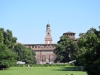 Château des Sforza à Milan avec murs en briques rouges, tour de l'horloge centrale et tours défensives. Pelouse verte au premier plan avec arbres.