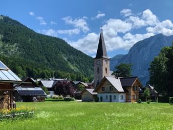 Village alpin d'Altaussee avec maisons traditionnelles, clocher et prairie verte entourés de montagnes boisées sous ciel bleu.