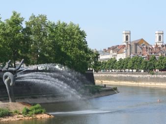 Sculpture de fontaine moderne pulvérisant de l'eau dans le Doubs à Besançon, avec des bâtiments historiques et deux clochers visibles de l'autre côté.