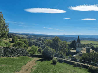 Vue panoramique sur le chemin de Stevenson montrant une chapelle blanche à dôme, collines verdoyantes, forêts et montagnes lointaines.