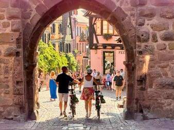 Deux cyclistes avec vélos traversent une arche en pierre vers une rue alsacienne colorée avec maisons à colombages et touristes.