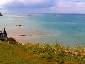 Vue panoramique d'Arromanches en Normandie avec falaises vertes, mer turquoise et vestiges des structures de débarquement du Jour J dans l'eau.