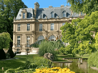 Manoir en pierre beige avec toit mansardé et fenêtres cintrées, entouré d'un jardin paysager avec étang, saules pleureurs et fleurs jaunes.