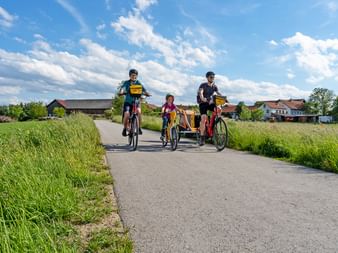 Famille de trois personnes à vélo sur route rurale dans le Salzkammergut avec enfant en remorque, entourée de champs verts et maisons.