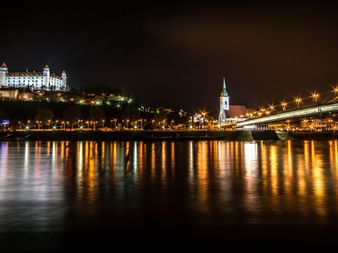 Vue nocturne du château illuminé de Bratislava sur une colline surplombant le Danube, avec un pont et les lumières de la ville.