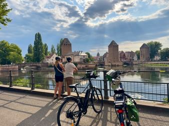 Deux cyclistes avec vélos admirant les tours médiévales des Ponts Couverts à Strasbourg sous un ciel nuageux dramatique.