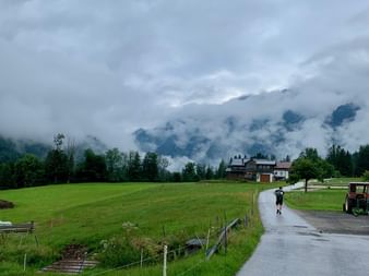 Route asphaltée humide traversant des prairies vertes au Salzkammergut avec maisons et montagnes couvertes de nuages sous un ciel nuageux.