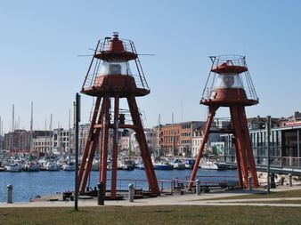 Deux balises de navigation rouge et blanc sur structures métalliques au port de Dunkerque. Marina avec voiliers et bâtiments portuaires en arrière-plan.