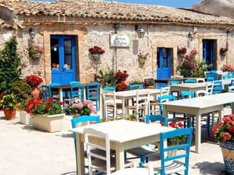 Terrasse de restaurant à Marzamemi avec tables blanches, chaises bleues et fleurs colorées. Bâtiment en pierre avec volets bleus et toit en terre cuite.