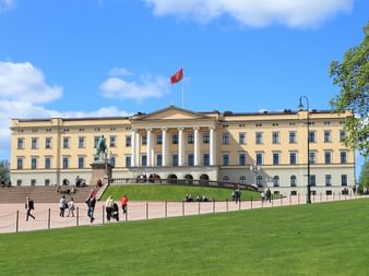 Palais royal d'Oslo, bâtiment néoclassique jaune avec drapeau norvégien. Pelouse verte au premier plan avec visiteurs et statue équestre.