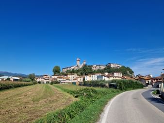 Village perché de Nozzano Castello avec clochers sous ciel bleu. Une route sinueuse traverse des champs verts vers le village historique.