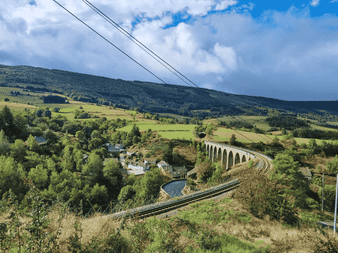 Viaduc ferroviaire en pierre traversant la campagne verdoyante du chemin Stevenson. Collines et petit village sous ciel bleu.