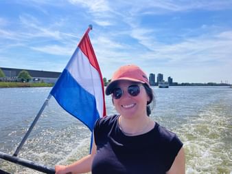Karen sourit sur un bateau tenant le drapeau néerlandais aux rayures rouge, blanc et bleu. Elle porte des lunettes de soleil et une casquette rose avec l'eau et la skyline derrière.