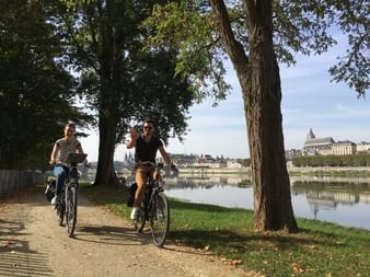 Deux cyclistes roulent le long de la Loire sur un chemin bordé d'arbres. Château historique visible de l'autre côté de l'eau avec pelouses.