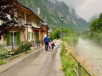 Adulte avec deux enfants marchant sur le sentier au bord du lac à Hallstatt, Autriche. Maisons traditionnelles bordent la rue avec montagnes brumeuses.