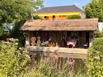 Ferme alsacienne miniature avec toit de tuiles au bord d'un étang. La structure ouverte montre un intérieur traditionnel avec figurines.