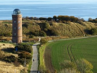 Phare en brique rouge avec lanterne en verre dans un paysage côtier. Champs verts et collines entourent la tour, cyclistes sur sentier proche.