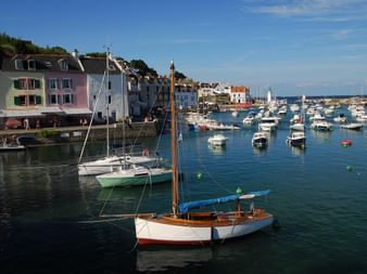 Port pittoresque de Belle-Île-en-Mer avec voiliers et bateaux à moteur amarrés dans une eau bleue calme, maisons colorées en bord de mer.