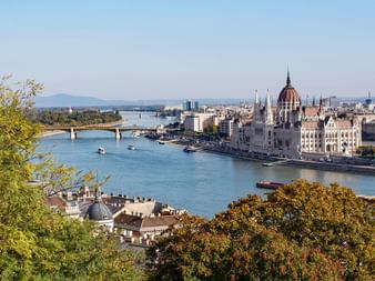 Vue de Budapest avec le Parlement hongrois le long du Danube, des ponts traversant l'eau et des arbres verts au premier plan.