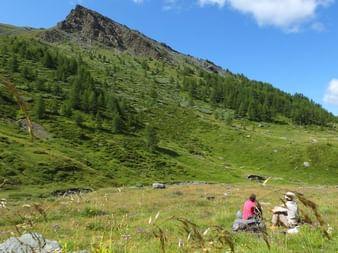 Deux randonneurs assis dans l'herbe dans la vallée du Queyras avec un pic rocheux spectaculaire au-dessus de pentes boisées vertes.