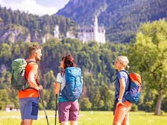 Randonneurs admirant le château Neuschwanstein Trois randonneurs avec sacs à dos debout dans un pré, regardant le château Neuschwanstein niché parmi les montagnes boisées de Bavière.