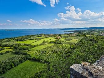 Vue panoramique du lac de Garde depuis Rocca di Manerba montrant les eaux bleues du lac, des champs verts, vignobles et collines sous un ciel nuageux.