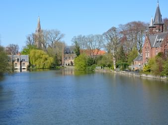 Vue pittoresque d'un lac à Bruges avec bâtiments historiques, clochers d'église et tour en brique rouge entourés d'arbres.